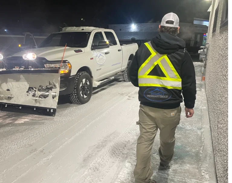 Worker in a reflective vest walking on a cleared, snow-covered sidewalk near a Summit Property Group truck equipped with a snowplow, showcasing professional snow removal services in Lethbridge.